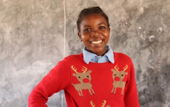 A child from Zambia smiling in school whilst wearing a red Christmas jumper.
